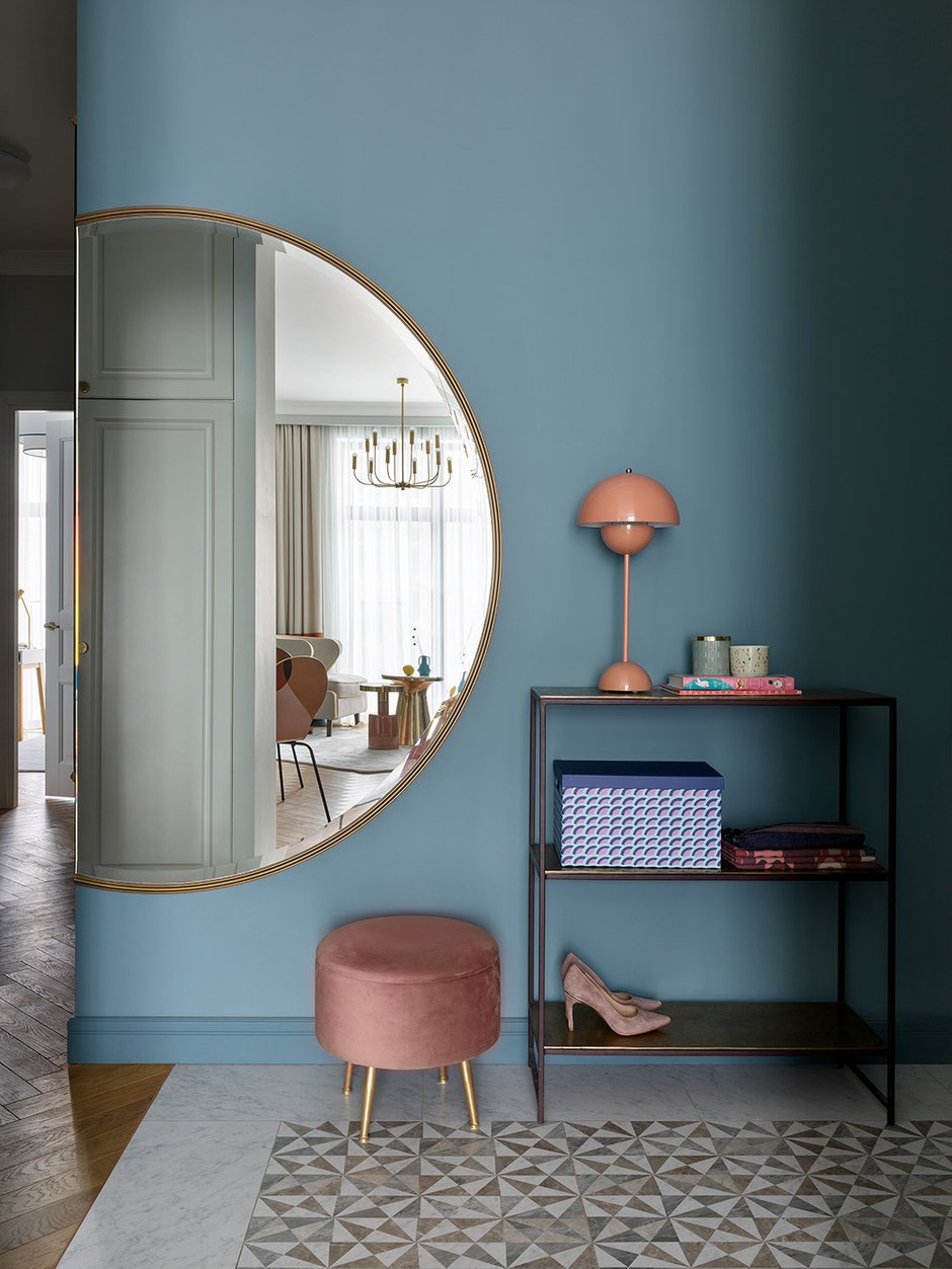 Для выделения зоны прихожей выбрали сложный темный синий цвет на одну стену. Это единственное место, где позволили себе темный тон. Но тут же задекорировали его частью круга большого зеркала, отражающего перспективу гостиной. Меньшая часть зеркала со смещением расположилась на продолжении стены под 90 градусов.