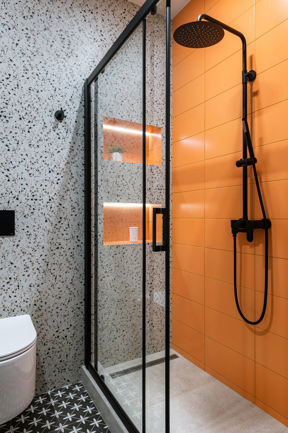 Основной материал в санузлах — крупноформатный керамогранит с рисунками под бетон и терраццо. В душевой акцентная плитка среднего размера, а на полу небольшая плитка с геометрическим рисунком. Смесители и фурнитура черного цвета, чтобы подчеркнуть графичность интерьера. Затирка эпоксидная, подобрана тон в тон к каждой плитке.