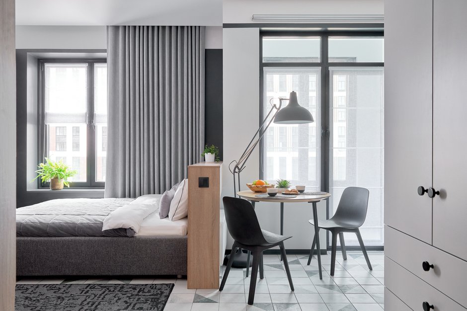 Окно у кровати оформлено легкими римскими шторами и портьерами с подкладкой блэкаут. В зоне кухни не использовали тяжелые портьеры, они бы смотрелись массивно.