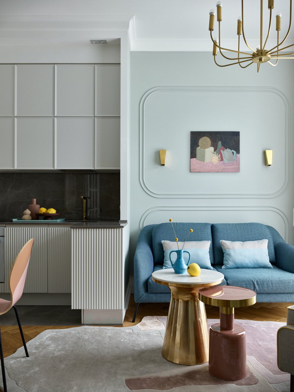 В гостиной установили небольшой, но прочный и симпатичный диванчик, сделанный по эскизам дизайнера, обитый в ткань «елочкой». Над ним — натюрморт модного современного художника Марка Якута, по цветам «собирает» все пространство гостиной и кухни. Гипсовые скругленные молдинги служат как бы рамой для неоформленной картины. Над креслом круглая полка, покрашенная вручную. Ковер от Art de Vivre необычной формы повторяет форму орнамента на стульях в столовой зоне. 