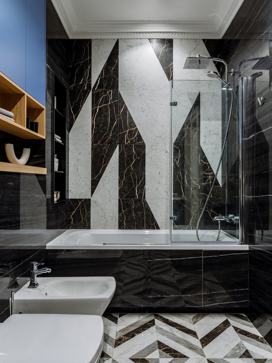 В отделке стен использовали крупноформатный керамогранит под мрамор и бетон. По широкой стороне ванны акцентное геометрическое панно из керамогранита. В отделке пола тоже использовали керамогранит с геометрическим рисунком. Строгие линии разбавили гипсовым карнизом.